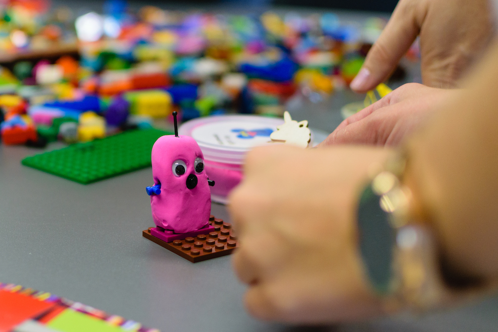 Photo ot table covered in lego with hands crafting a pink play doh monster