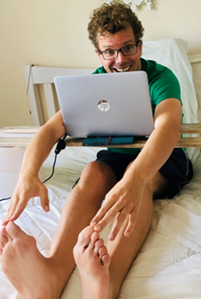Man on a bed with a laptop, smiling and touching his toes